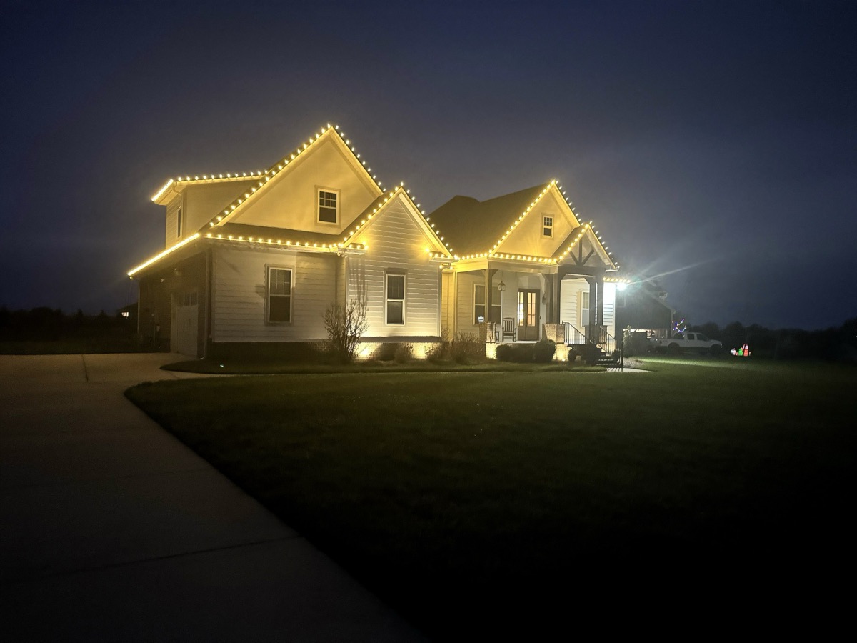 White farmhouse at dusk with warm white lights outlining every gable and porch