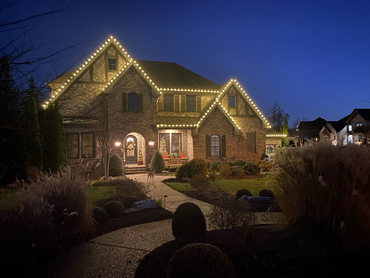 Brick home at twilight outlined in warm white lights along each roof peak